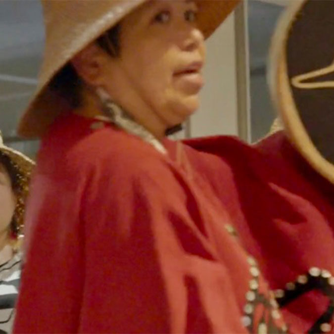 haisla-heritage-our-canoe-song Person in traditional attire, wearing a wide-brimmed hat and holding an item, stands indoors with another similarly dressed person partially visible behind.
