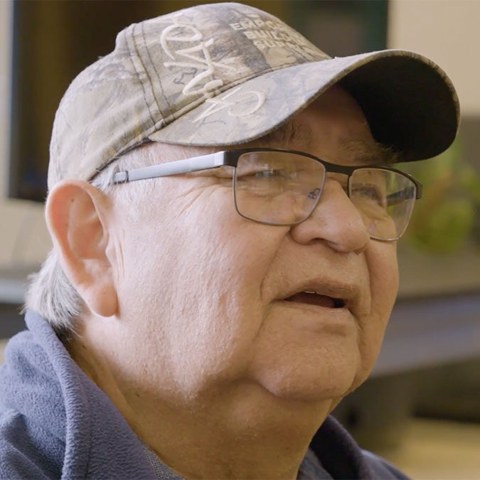 An elderly man wearing glasses and a camo baseball cap, smiling slightly.