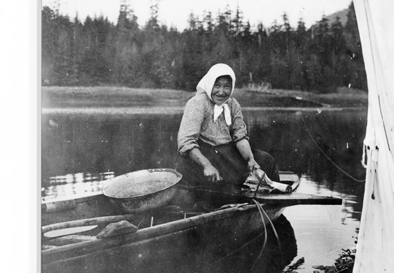 A smiling woman with a headscarf sits on a wooden structure over a lake, preparing fish, with a forested shoreline in the background.