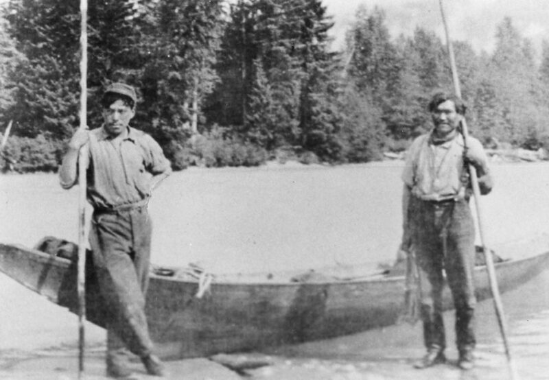 Two men standing on the shore next to a wooden boat with oars, surrounded by trees in the background.