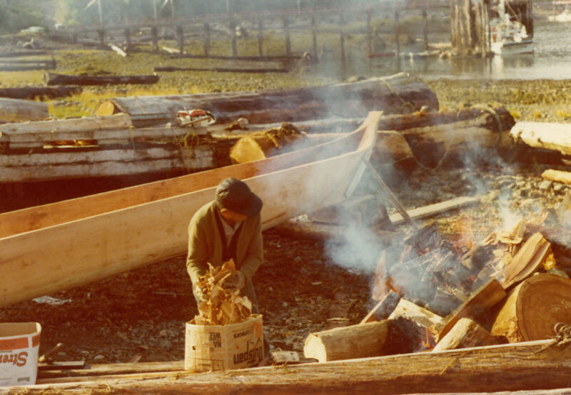 A person stands near a fire and wooden logs, working outdoors by the water with trees and structures visible in the background.