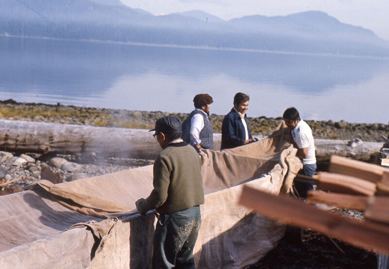 Four people, three men and one woman, work together by a large cloth near a calm lake with mountains in the background.