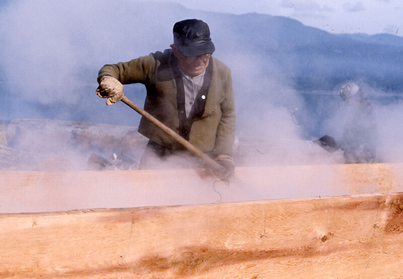 A person wearing gloves and a hat uses a tool to manipulate steaming wood outdoors amid foggy conditions.