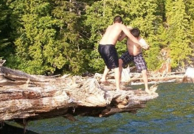 Two shirtless people, one adult and one child, balance on a fallen tree over water with trees in the background.