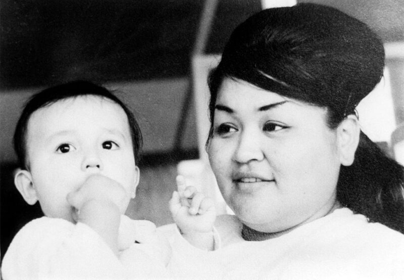A woman holds a baby who is sucking on their hand. Both are looking in the same direction with a neutral expression. Black and white photo.
