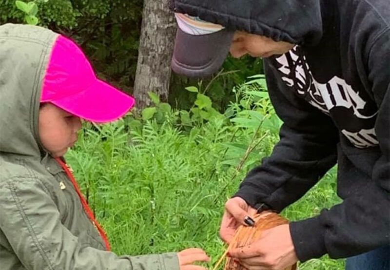Two people outdoors, one child in a green jacket and pink hat, the other in a black hoodie, working together to weave or tie something with natural materials.