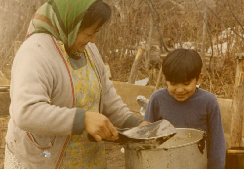 A woman in a headscarf and apron stirs a large pot outdoors while a young boy watching smiles.