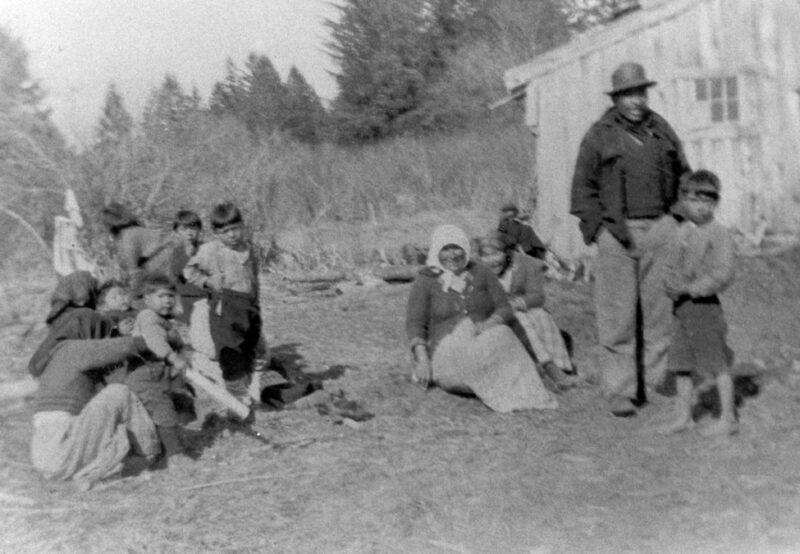 A group of people, including adults and children, are gathered outdoors near a wooden building in a rural area. Some are standing, and others are seated.