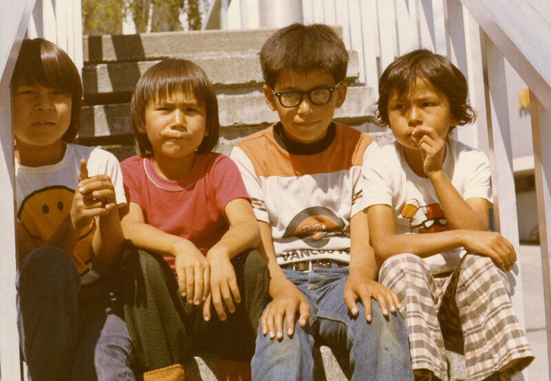 Four children sit on outdoor steps, three boys and one girl, with one boy holding a banana. All are casually dressed, wearing t-shirts and pants.