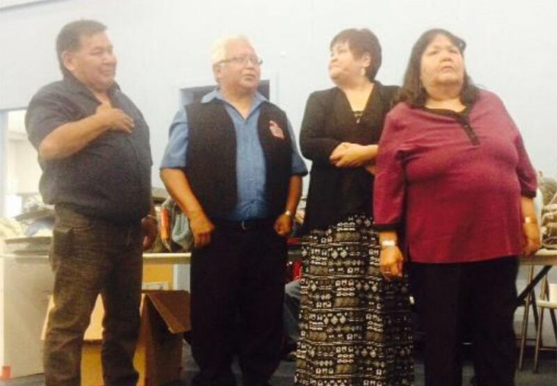 Four people standing together indoors, with one person placing their hand on their chest, and the others standing with neutral expressions.