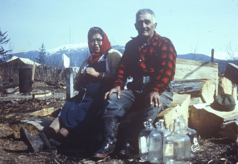 An older man and woman sit outdoors on logs with chopped wood and glass bottles around them, with snowy mountains in the background.
