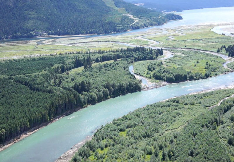 Aerial view of a winding river through dense green forests and fields, leading to a larger body of water in the background, surrounded by hilly terrain.