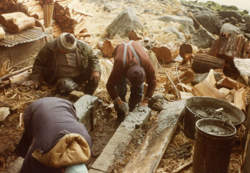 Three workers in warm clothes, kneeling and working with concrete near a pile of firewood in an outdoor setting with rocks and straw on the ground.