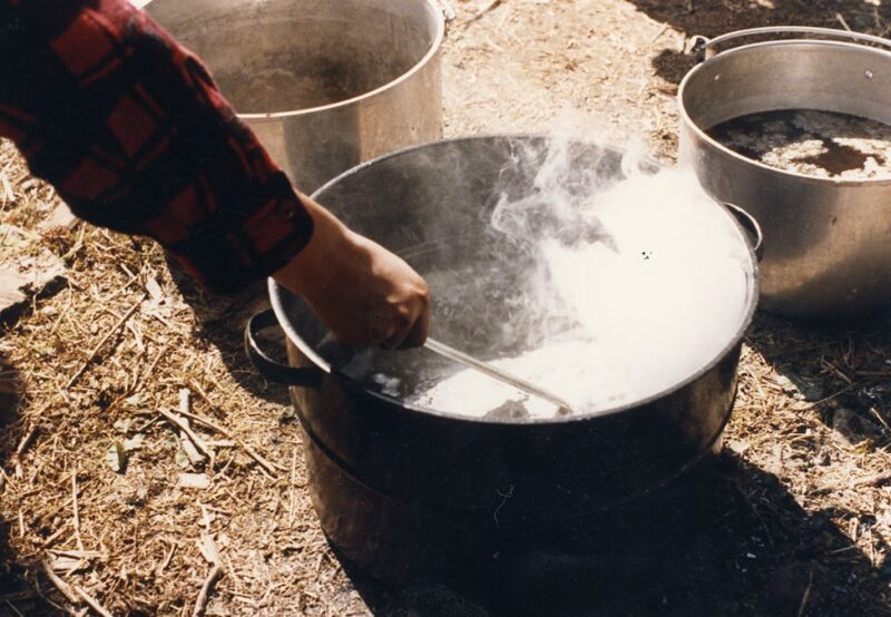 Person stirring bubbling liquid in a black pot using a metal spoon outdoors, with two other pots nearby on a dirt surface.