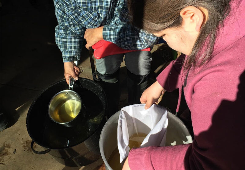 Two people filtering liquid into a pot using a white cloth; one holds a container, the other holds the cloth.